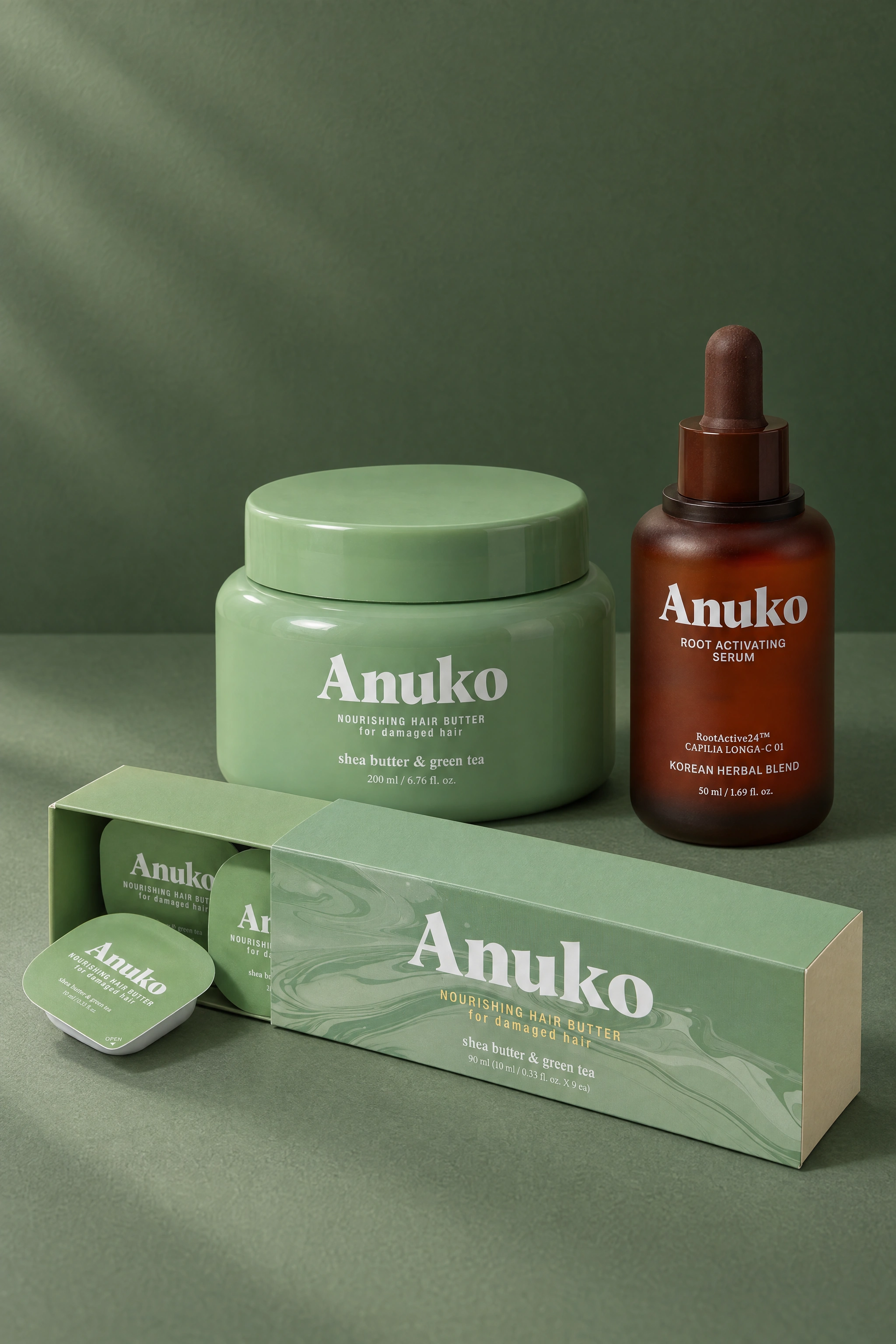 Anuko Nourishing Hair Butter jar and Travel Hair Butter carton beside the Root Activating Serum bottle on a sage studio ground